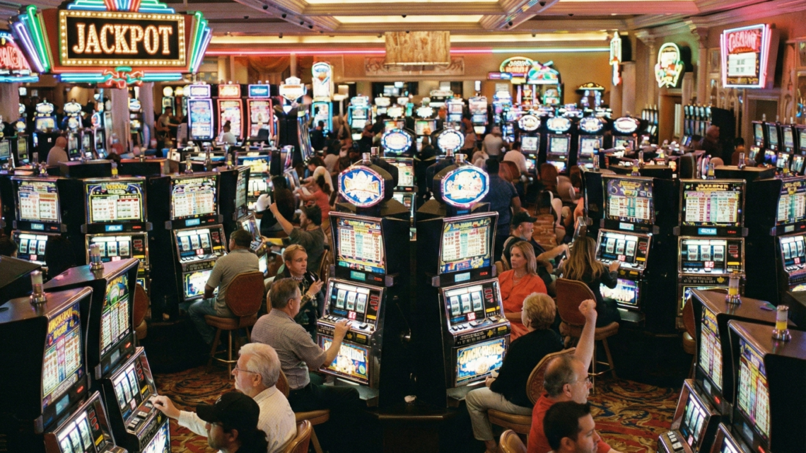 Pemandangan kasino dengan mesin slot yang ramai, para pemain sedang memainkan mesin slot di lantai kasino dengan pencahayaan warna-warni yang terang.